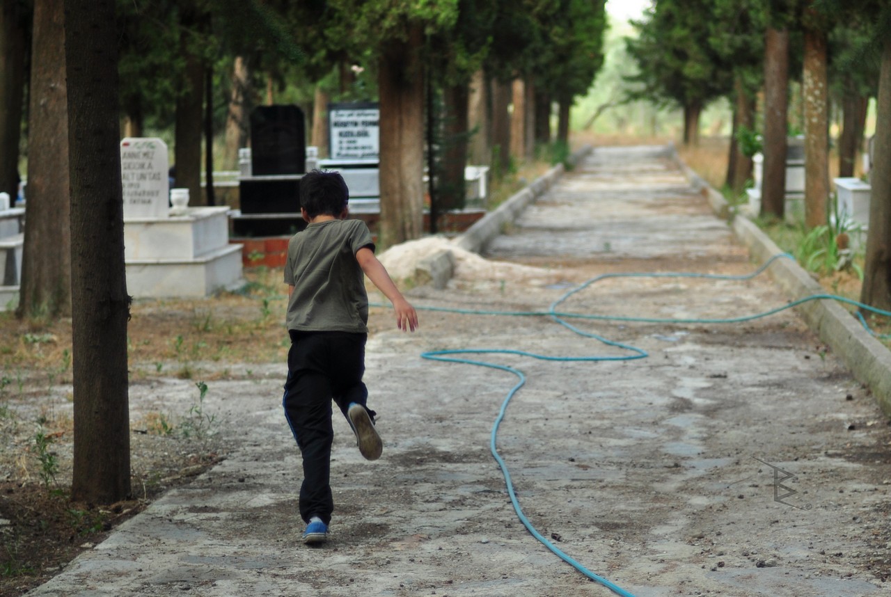 A child running along a tree-lined cemetery path with headstones and a blue hose.