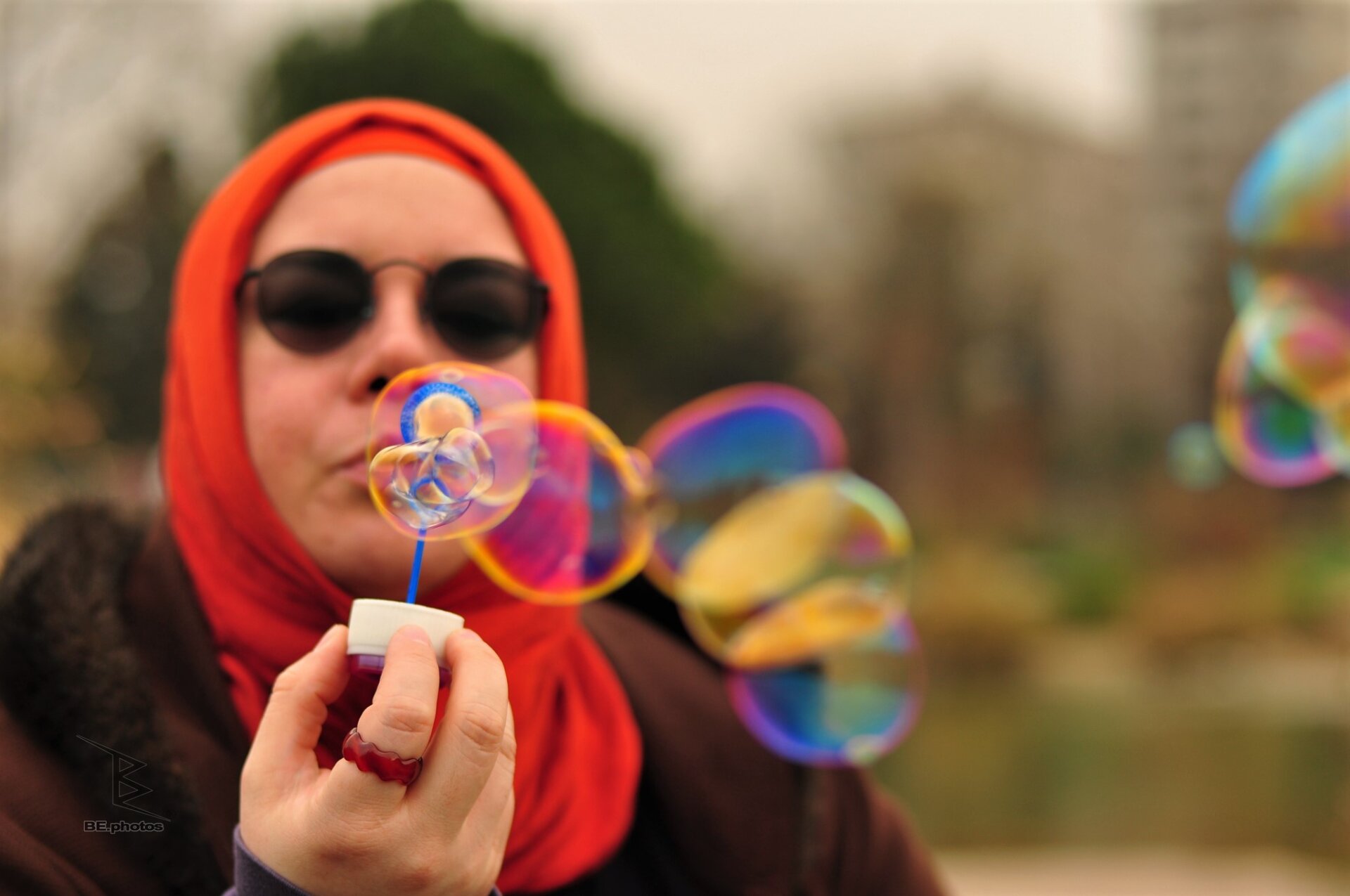 Woman in a red headscarf blowing a stream of colorful soap bubbles outdoors.