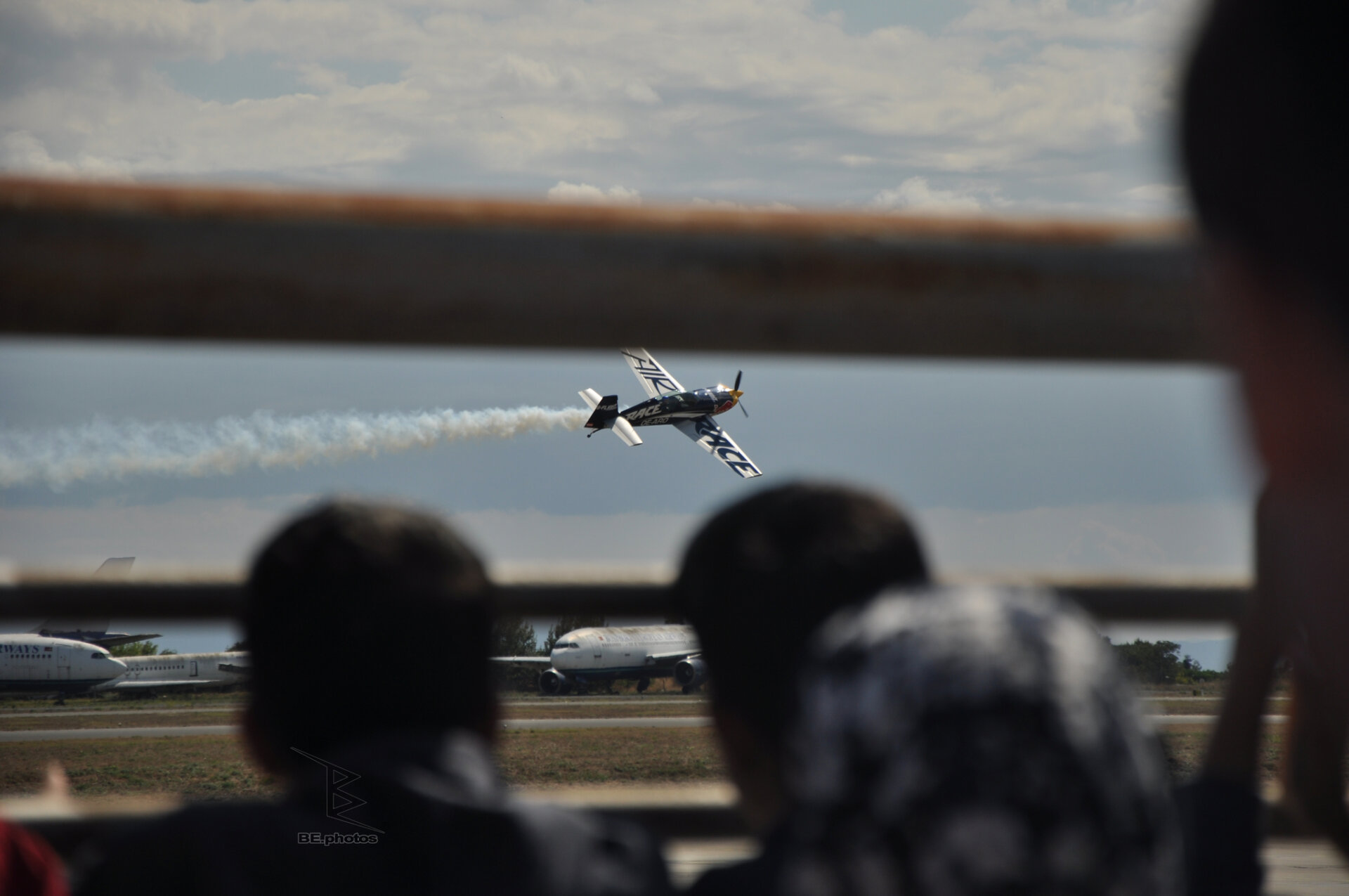 A Red Bull Air Race plane crossing the sky with a smoke trail, blurred spectators and parked airliners below.
